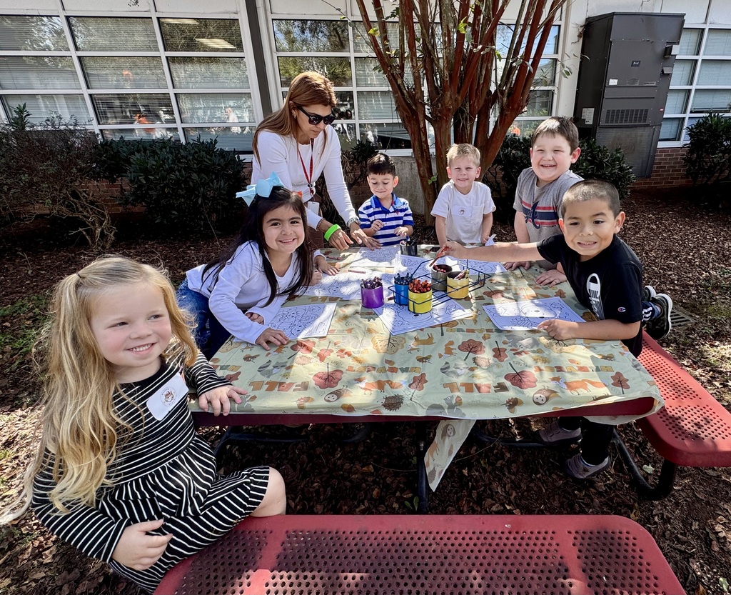 A group of young students and an adult supervisor gather around an outdoor picnic table coloring fall-themed activity sheets with crayons.