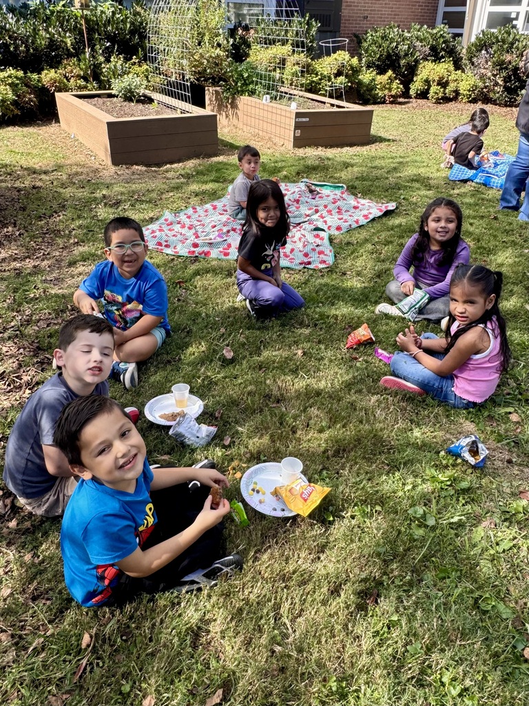 A A group of young elementary students enjoying an outdoor picnic snack time on a sunny grassy courtyard near raised garden beds. The children are seated on the grass and on a red floral picnic blanket, eating snacks from paper plates and baggies while smiling at the camera. In the foreground, a young boy in a blue shirt grins as he holds a snack next to his paper plate and cup.