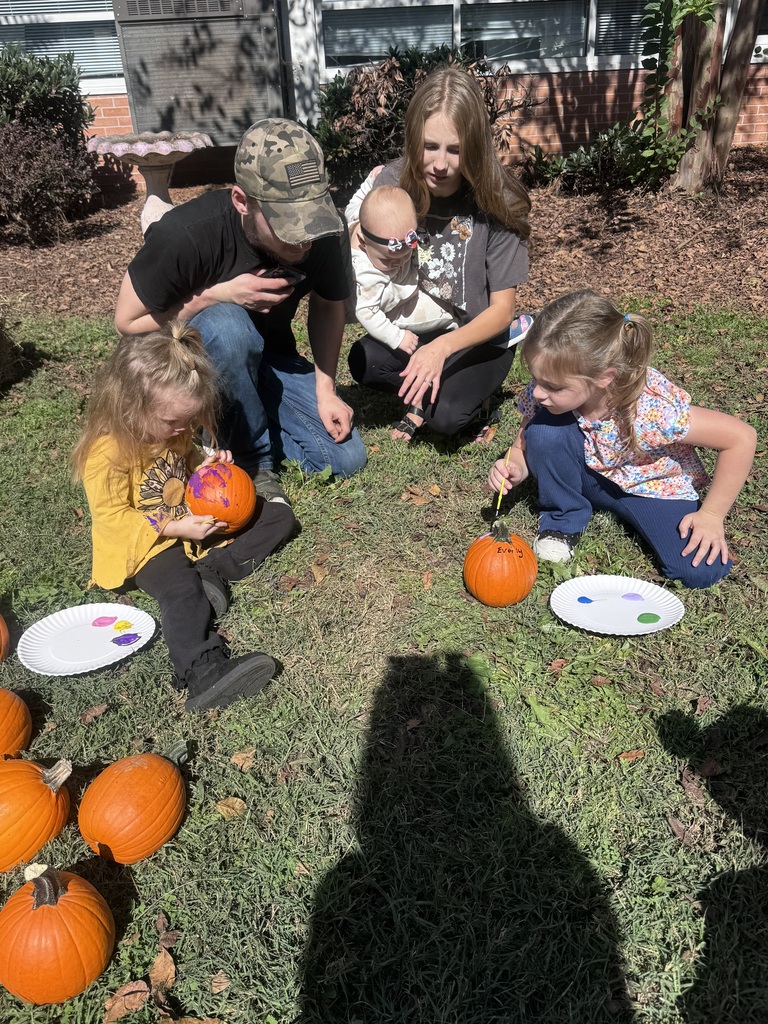 A family sits together outdoors on a grassy lawn during a sunny pumpkin painting activity. Two young girls crouch on the grass, using paintbrushes to paint small orange pumpkins resting on paper plates with spots of colorful paint. The girl on the left wears a long-sleeved yellow sunflower shirt, and the girl on the right wears a floral top and blue pants. A man wearing a camouflage cap and a woman holding a baby kneel behind them, watching intently.