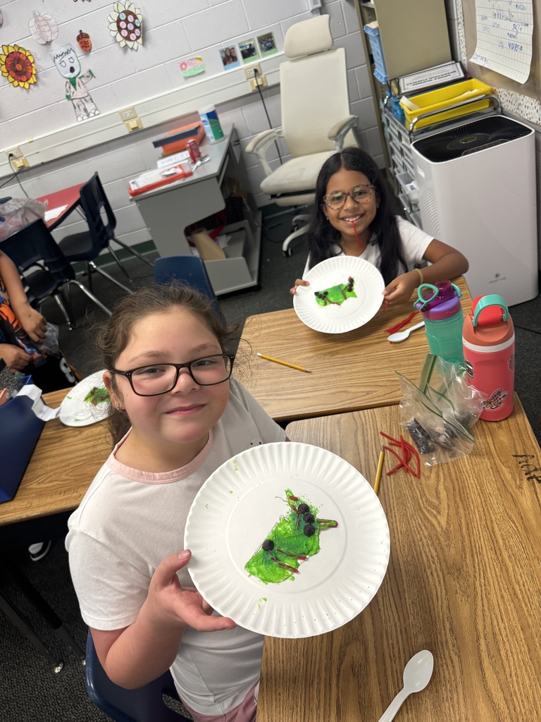 Two elementary school girls wearing glasses sit at wooden classroom desks, smiling as they present paper plates decorated with green frosting caterpillars made during a classroom activity.
