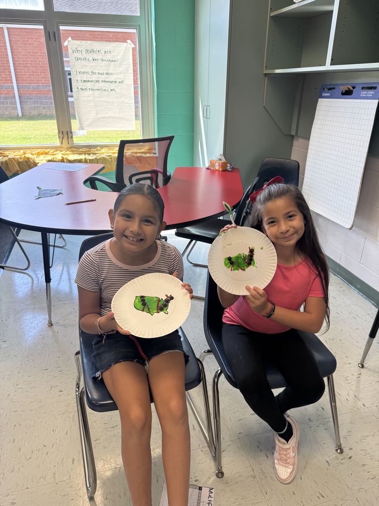 Two elementary school girls sit side-by-side in blue chairs near a horseshoe-shaped table, holding up paper plates featuring green caterpillar crafts created with frosting, licorice, and candy pieces.