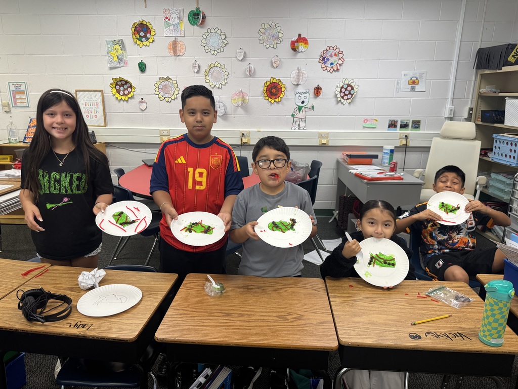 Five smiling elementary students stand and sit behind classroom desks, holding up paper plates featuring edible caterpillar crafts made with green frosting, candy toppings, and red licorice. Autumn flower decorations are displayed on the white cinder block wall behind them.