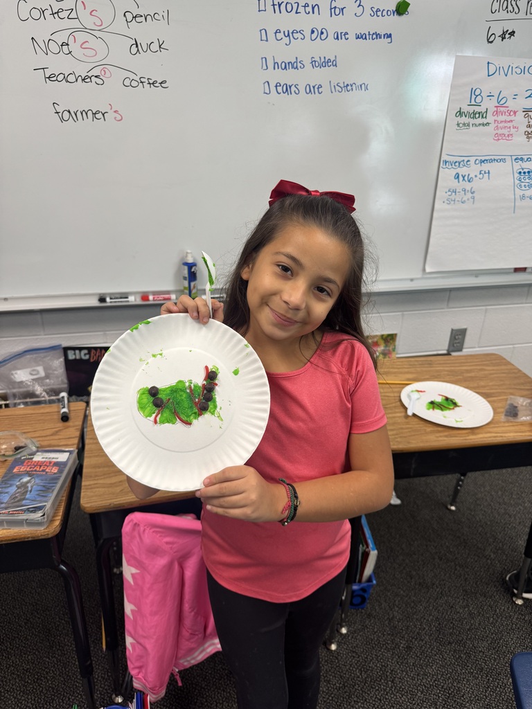 A young girl with a red bow in her hair stands next to a classroom whiteboard, holding up a paper plate featuring a green caterpillar craft made with green frosting, red licorice legs, and chocolate chip details.