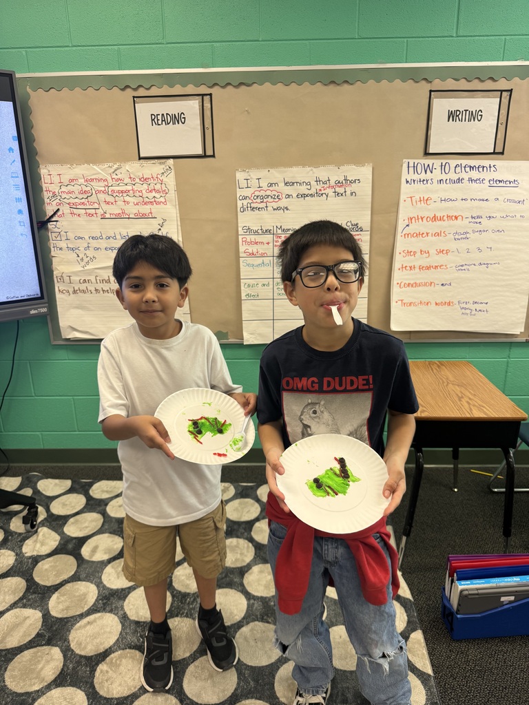 Two elementary school boys stand side-by-side in front of classroom anchor charts titled "READING" and "WRITING," each holding up a paper plate displaying a caterpillar craft decorated with green frosting, chocolate chips, and red licorice.