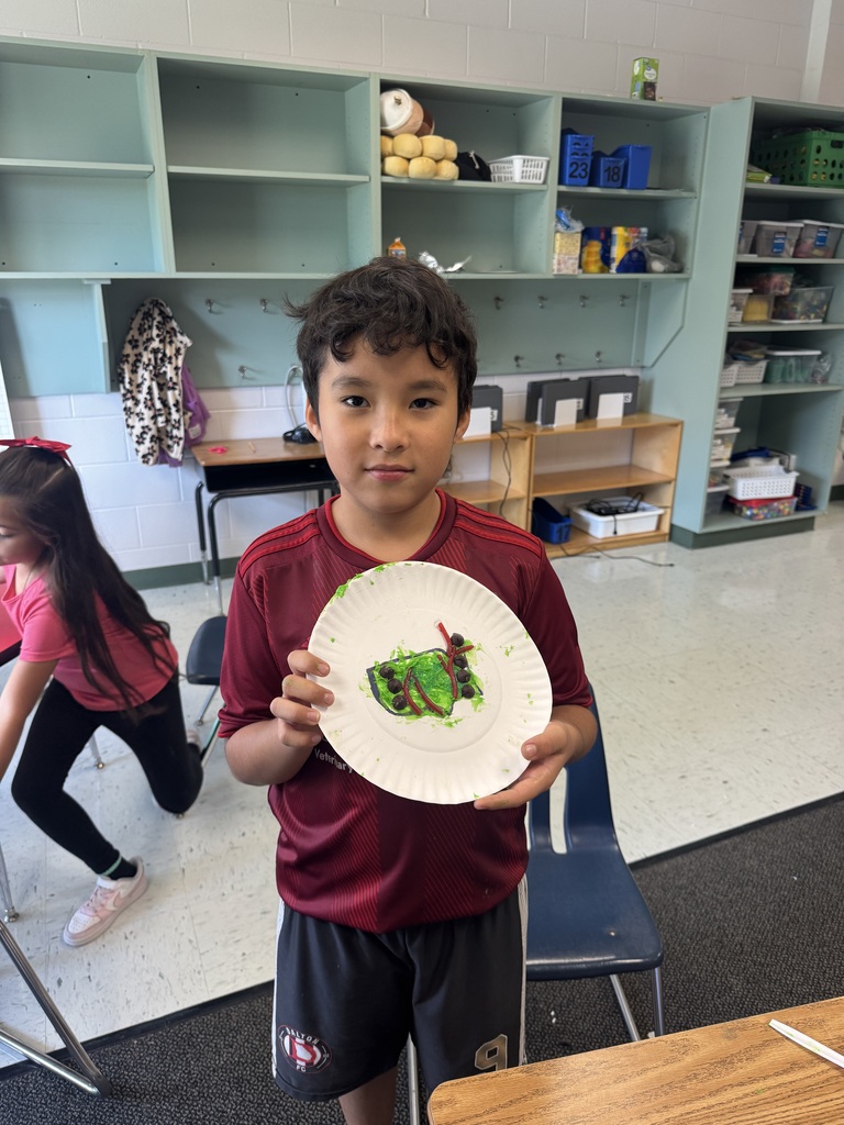A young boy wearing a maroon Dalton FC jersey stands in a classroom, holding out a paper plate decorated with a green icing caterpillar craft. Classmates and classroom shelving units are visible in the background.