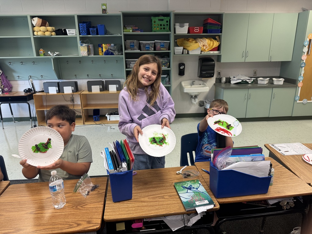 Long Alt Text: Three elementary students sit at wooden desks in a classroom, proudly displaying paper plates decorated with green icing, red licorice strands, and chocolate chips to look like caterpillars. The classroom background features green storage cabinets, shelves, and a paper towel dispenser.Long Alt Text: Three elementary students sit at wooden desks in a classroom, proudly displaying paper plates decorated with green icing, red licorice strands, and chocolate chips to look like caterpillars. The classroom background features green storage cabinets, shelves, and a paper towel dispenser.