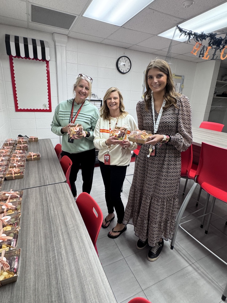 Three women standing together in a breakroom or classroom, each holding a small individual charcuterie box wrapped with a bow. On the left, long grey tables are lined with numerous matching charcuterie gift boxes. The room features red chairs, white walls, and Halloween banner decorations hanging from the ceiling.
