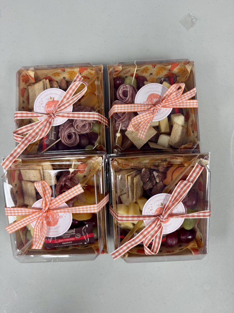 Four clear square plastic gift boxes arranged in a two-by-two grid on a light grey surface. Each box contains an individual mini charcuterie board featuring autumn-themed tissue paper lining. Inside the boxes are assorted items including rolled salami, cheese cubes, crackers, grapes, dried apricots, nuts, and a packaged Bouchard dark chocolate piece. Every box is tied with an orange-and-white gingham ribbon finished with a bow and features a round sticker reading "Thank You!" with fall-themed text and pumpkin illustrations.
