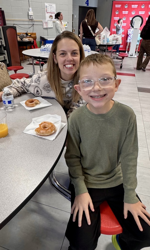 A smiling adult woman and a young boy wearing glasses and braces sit together at a cafeteria table enjoying donuts and juice during a school breakfast event.