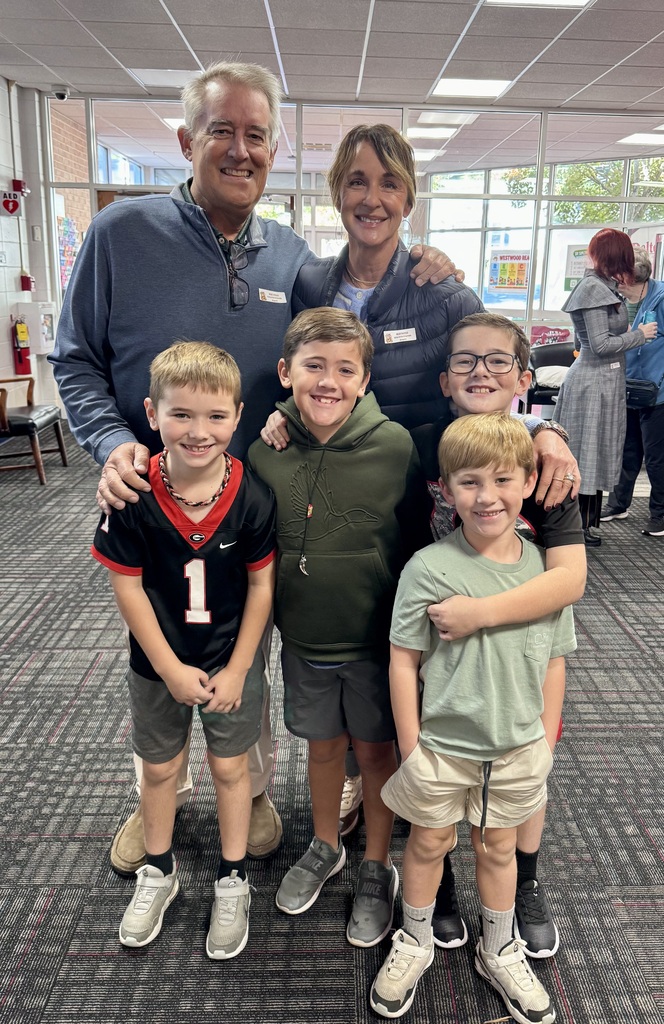An adult couple stands behind four smiling young boys posing together in a school hallway. One boy wears a Georgia Bulldogs jersey, while another wears a green hoodie.