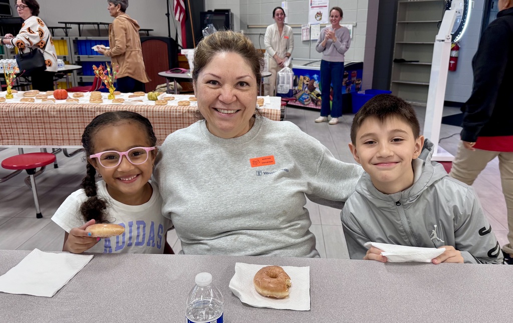 An adult woman sits at a school cafeteria table between two smiling elementary students—a young girl wearing pink glasses and an Adidas shirt on the left holding a donut, and a boy in a grey jacket on the right. Plates with donuts and a bottle of water sit on the table in front of them.
