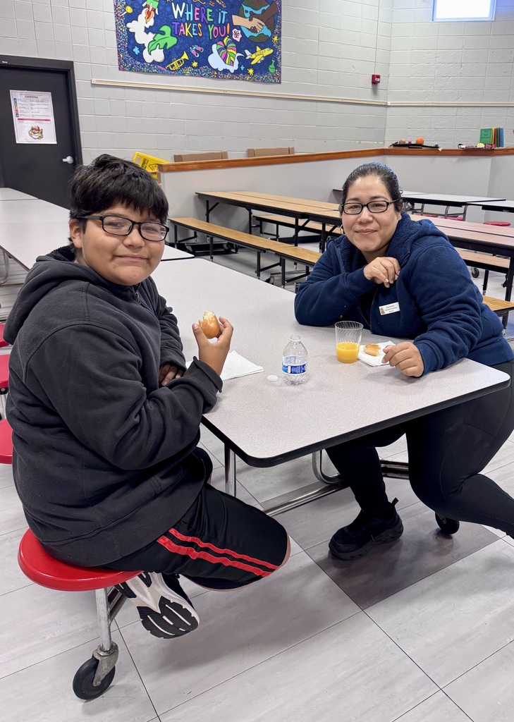 A young student wearing glasses and a grey hoodie sits at a cafeteria table enjoying a donut and juice alongside an adult woman wearing glasses and a blue jacket.