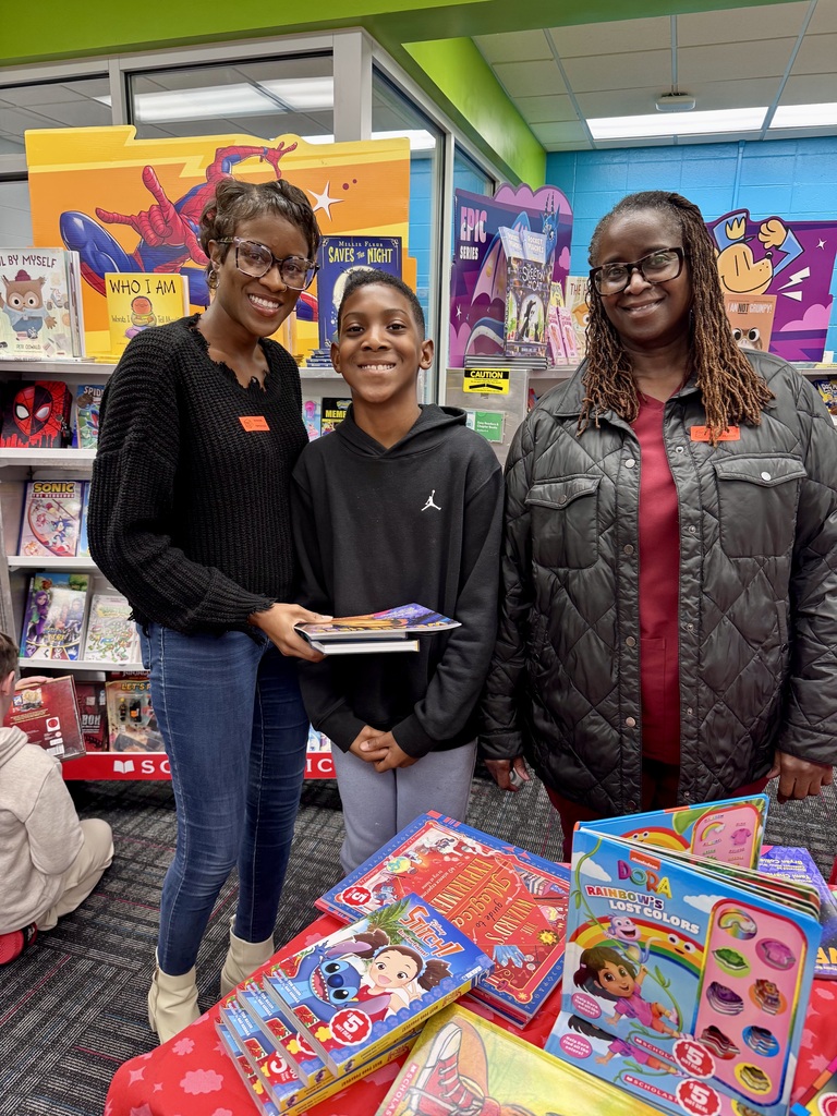 Two women stand on either side of a middle school student in a school book fair setting filled with colorful books. The boy holds a stack of new books while all three smile at the camera.