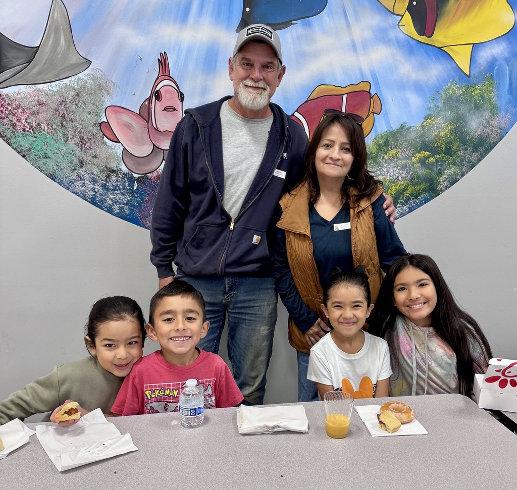A family of six poses together at a school cafeteria table beneath a large ocean mural. Two adults stand behind four young children seated at the table with donuts and juice in front of them.