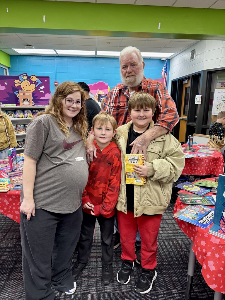A pregnant woman, a bearded older man, and two young boys pose together in a school library setting filled with book displays. The older boy holds a book while the group smiles for the camera.