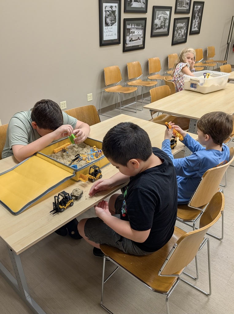 Several children sit at low wooden tables playing with sand and small toy construction vehicles, while a girl stands nearby next to a bin of rice.