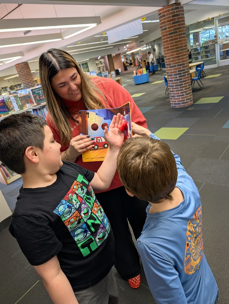 An adult woman in a red sweatshirt holds an open book while two boys look on, with one boy pointing at the page inside a library with brick pillars.