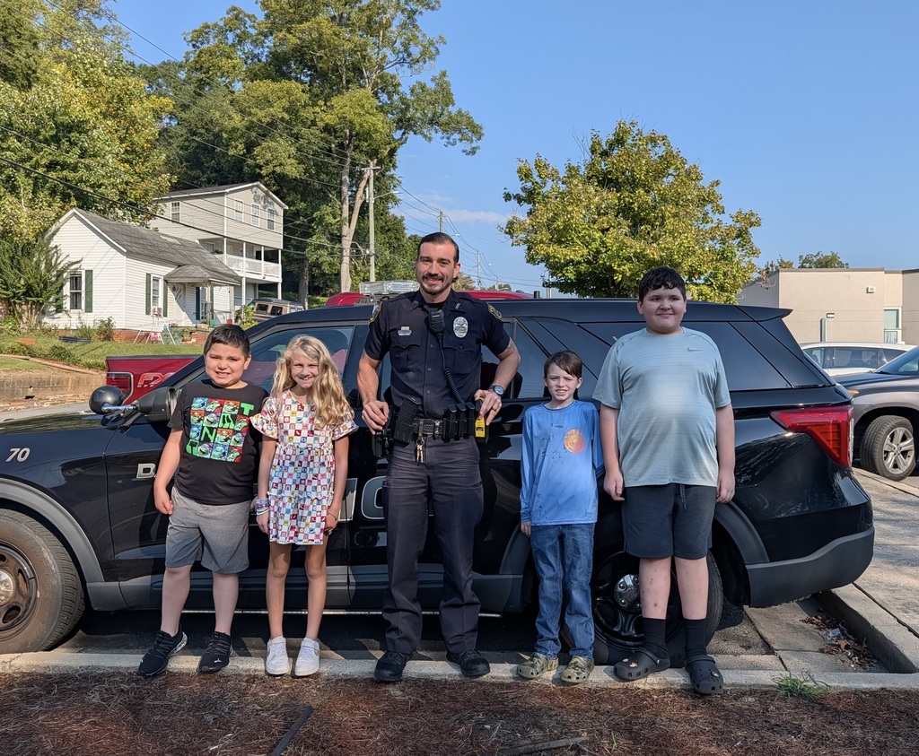 A police officer in uniform stands centered in front of a black police SUV, flanked by four smiling children posing outdoors on a sunny day.
