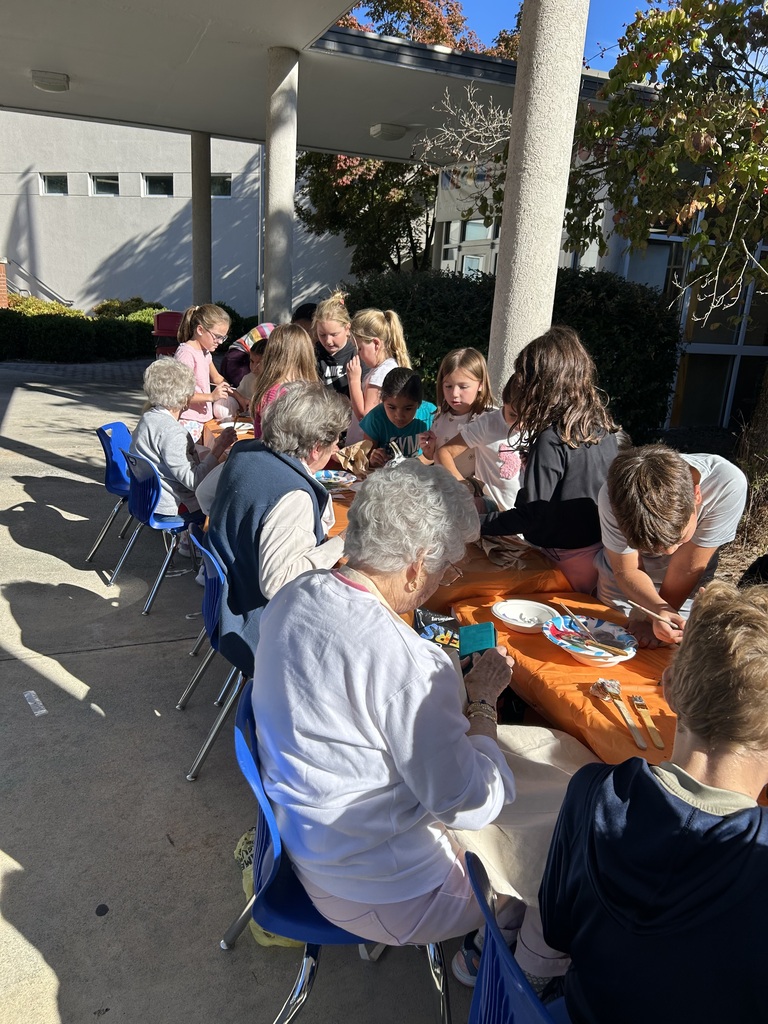 An angled view of children and senior citizens sitting at long, orange-covered tables outdoors, painting pumpkins together.