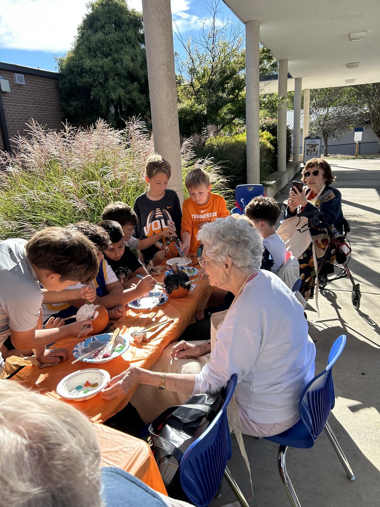 Children and senior adults gathered at outdoor tables under a portico, working together on a pumpkin-painting activity.