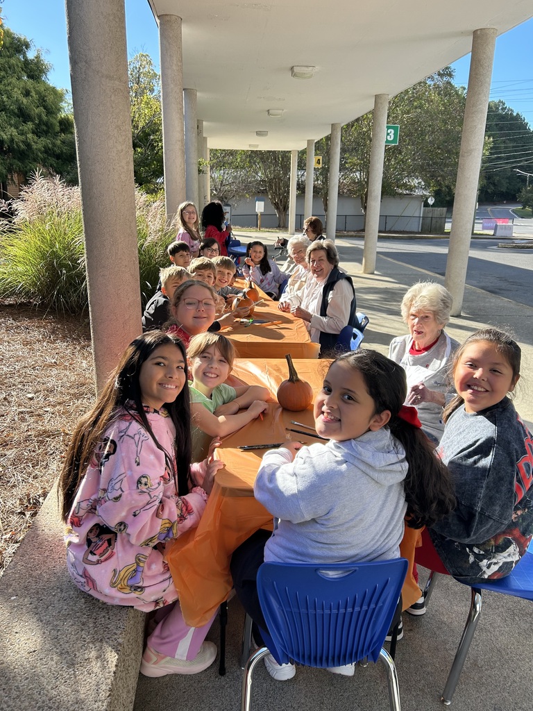 Elementary students and senior volunteers sitting together at an outdoor table covered with an orange cloth, painting pumpkins under a covered walkway.