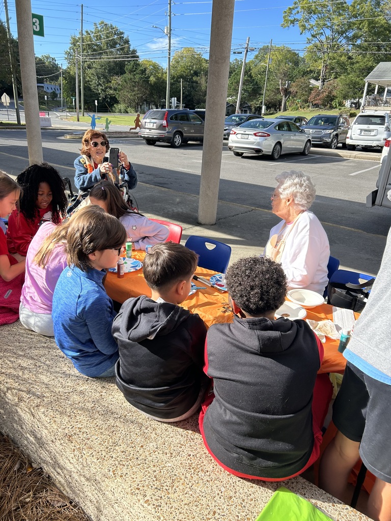 Students and volunteers seated along outdoor tables under a breezeway painting small pumpkins, with parked cars visible on the street in the background.