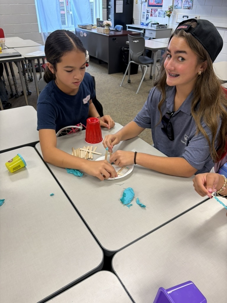 Two girls sitting at a classroom desk building a structure on a paper plate with craft sticks, blue play dough, and a red plastic cup.