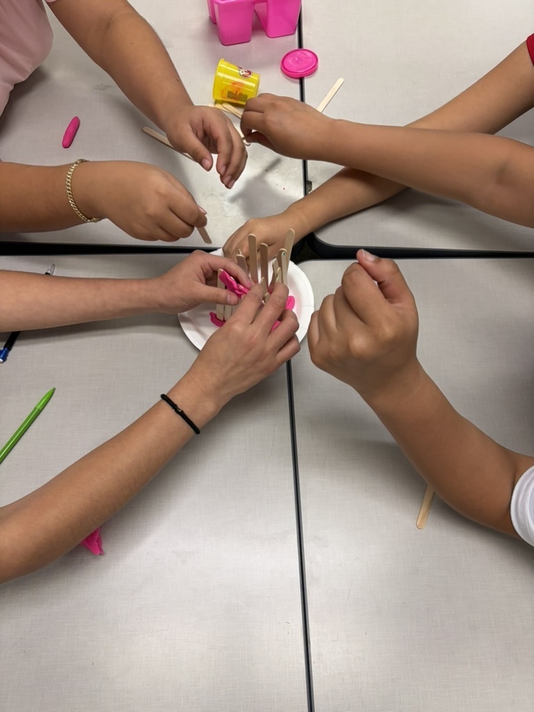 Overhead shot of several students' hands working together to arrange craft sticks upright in pink play dough on a paper plate.