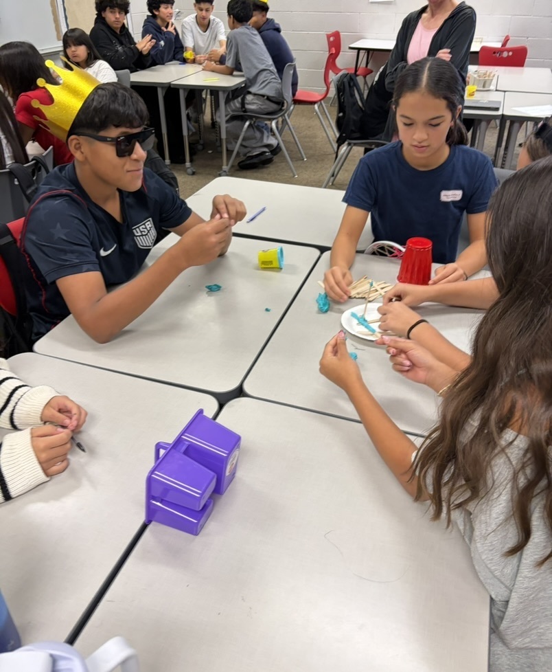 A group of students, including a boy wearing sunglasses and a gold paper crown, sitting at classroom desks building with craft sticks and play dough.
