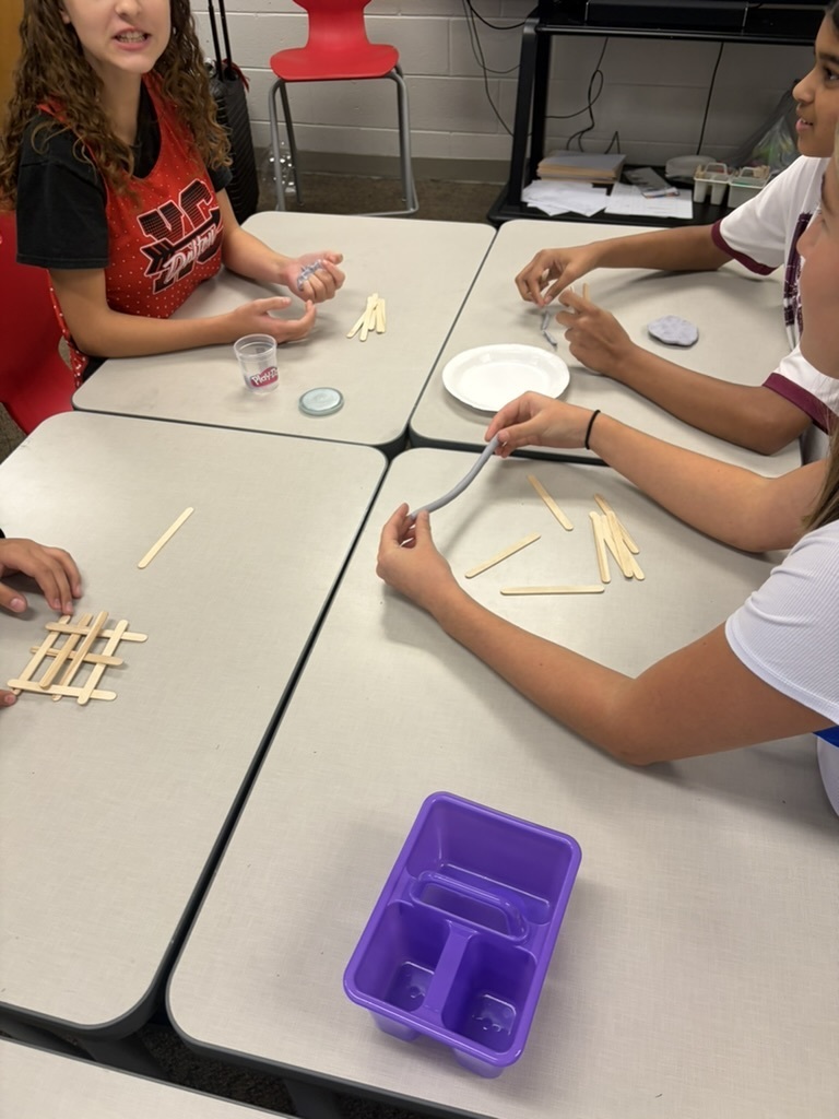 Students sitting at desks working with gray modeling clay and craft sticks, with one student shaping a clay rope and another assembling a stick grid.
