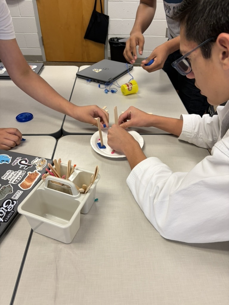 Students working at a classroom desk using blue modeling clay and craft sticks to construct an engineering project on a paper plate.