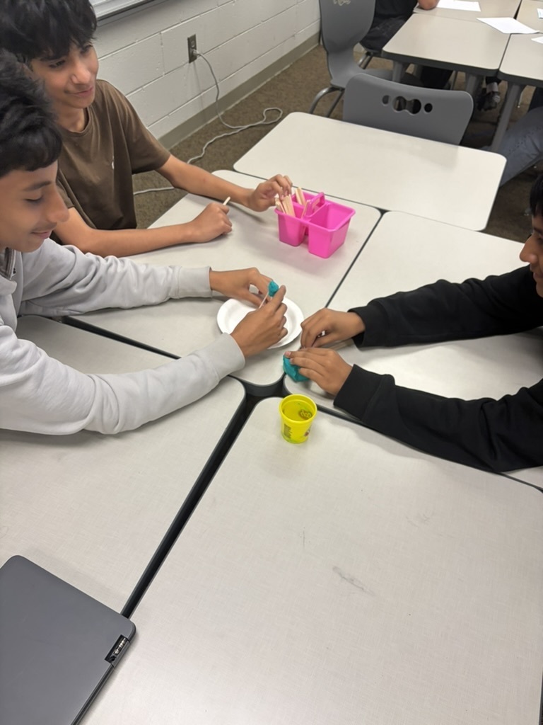 Three students sitting at desks collaborating on a STEM activity using craft sticks, a paper plate, and teal play dough.