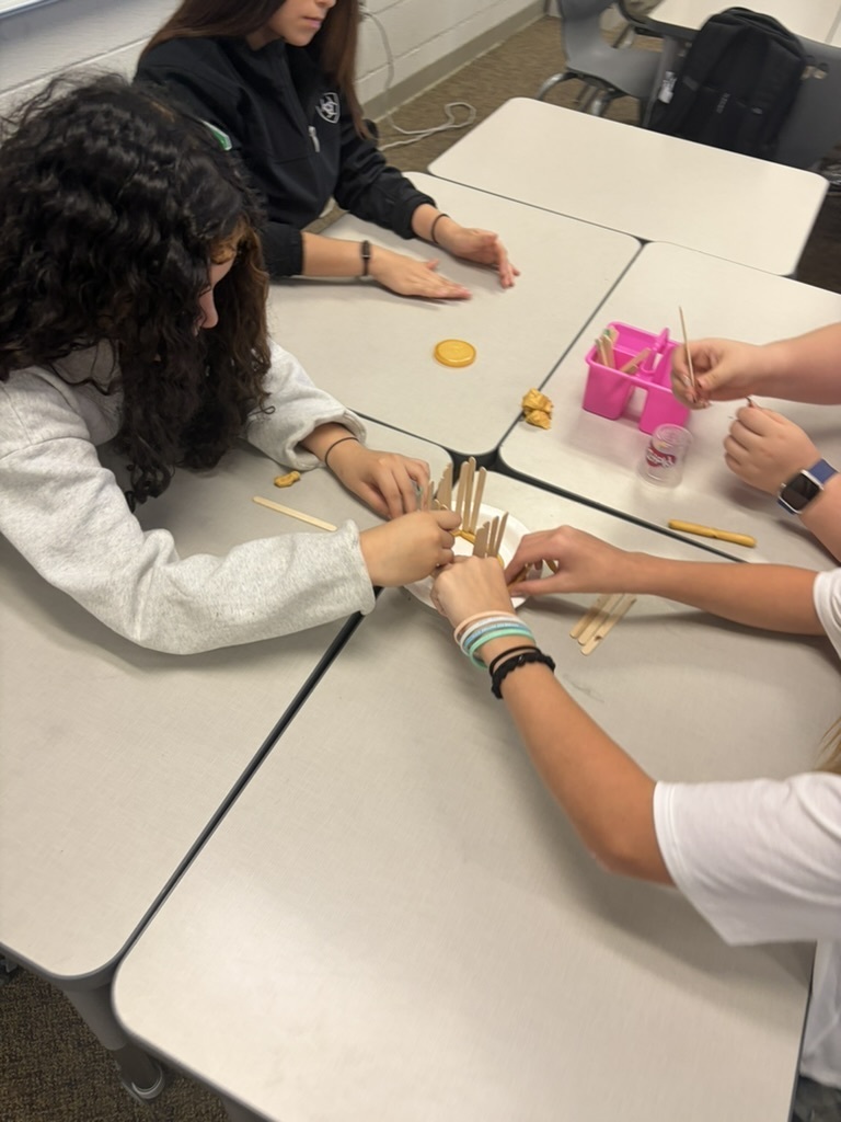 Students gathered around a desk placing craft sticks vertically into yellow play dough on a paper plate during a group challenge.