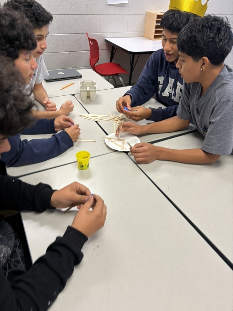 A group of boys sitting around joined desks collaborating on a craft stick and play dough building activity.