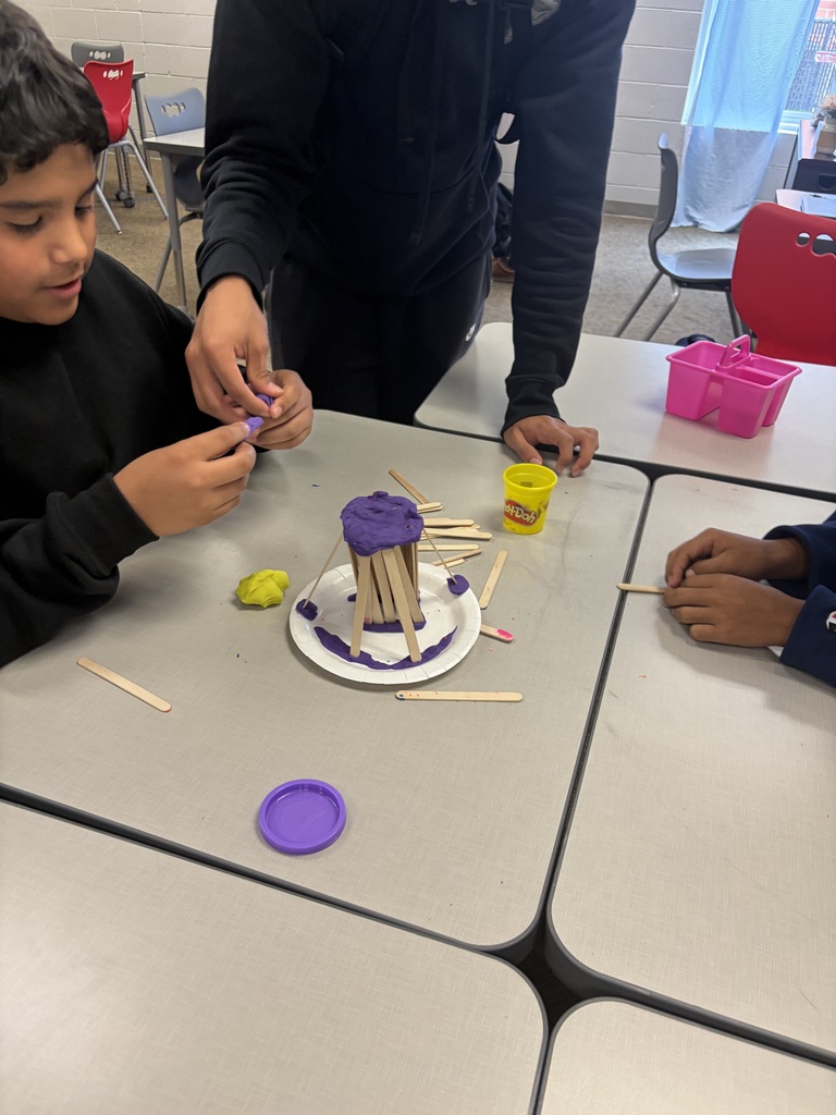 Middle school students seated around a desk building a tower structure using craft sticks, purple play dough, and a paper plate.