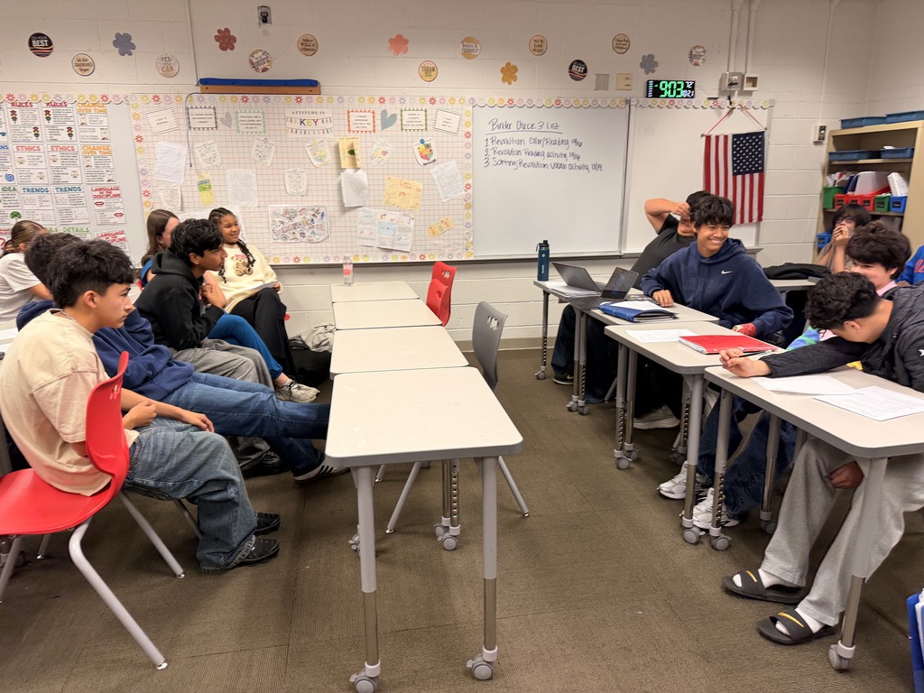 Students smiling and engaging with each other while seated at facing desks during a classroom debate session.