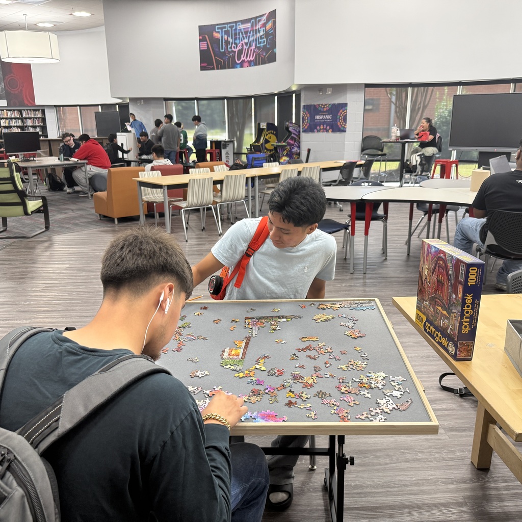 Two students sitting across from each other in a school media center working on a 1,000-piece puzzle on a dedicated puzzle board.