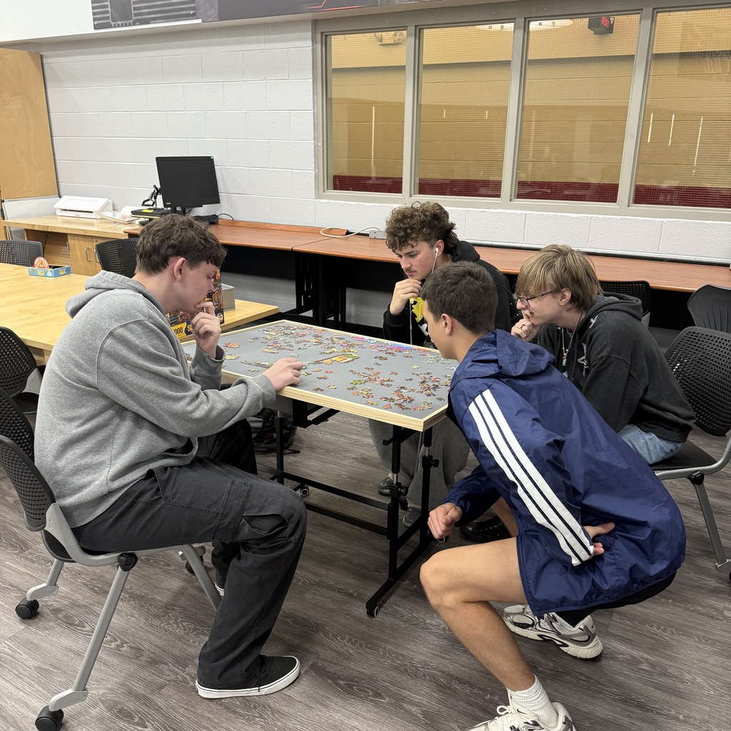 Four teenage boys gathered closely around a table in a library focusing on putting together a large jigsaw puzzle.