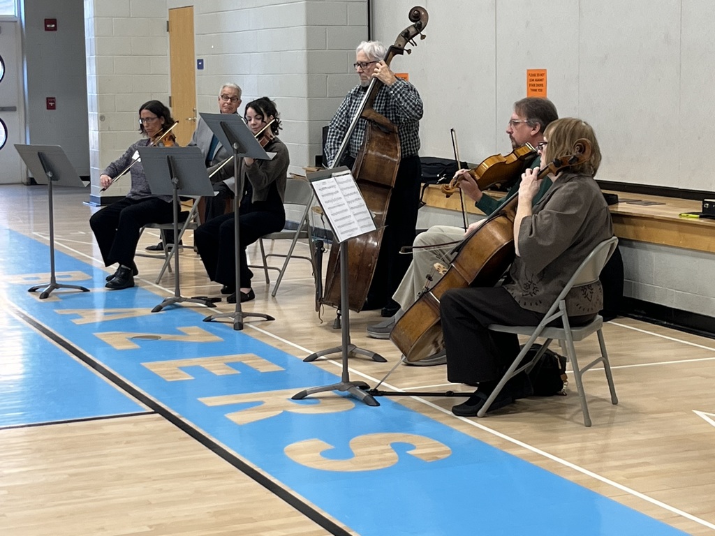 A small orchestra ensemble performs inside the Intermediate Center gym. Five musicians play string instruments—violins, cello, and bass—seated in a semicircle in front of music stands. The scene shows the bright blue “BLAZERS” lettering on the gym floor behind them.