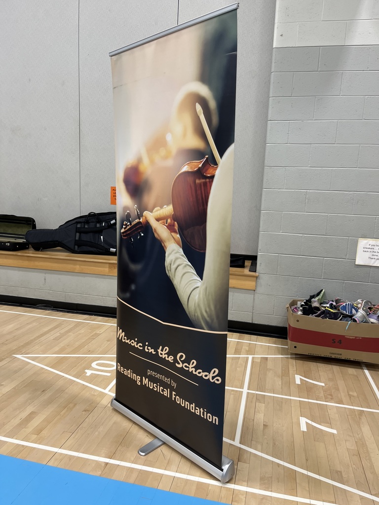 A banner reading “Music in the Schools presented by Reading Musical Foundation” stands in the gym. The banner features a close-up image of musicians playing string instruments, set against the gym’s hardwood floor and gray wall.
