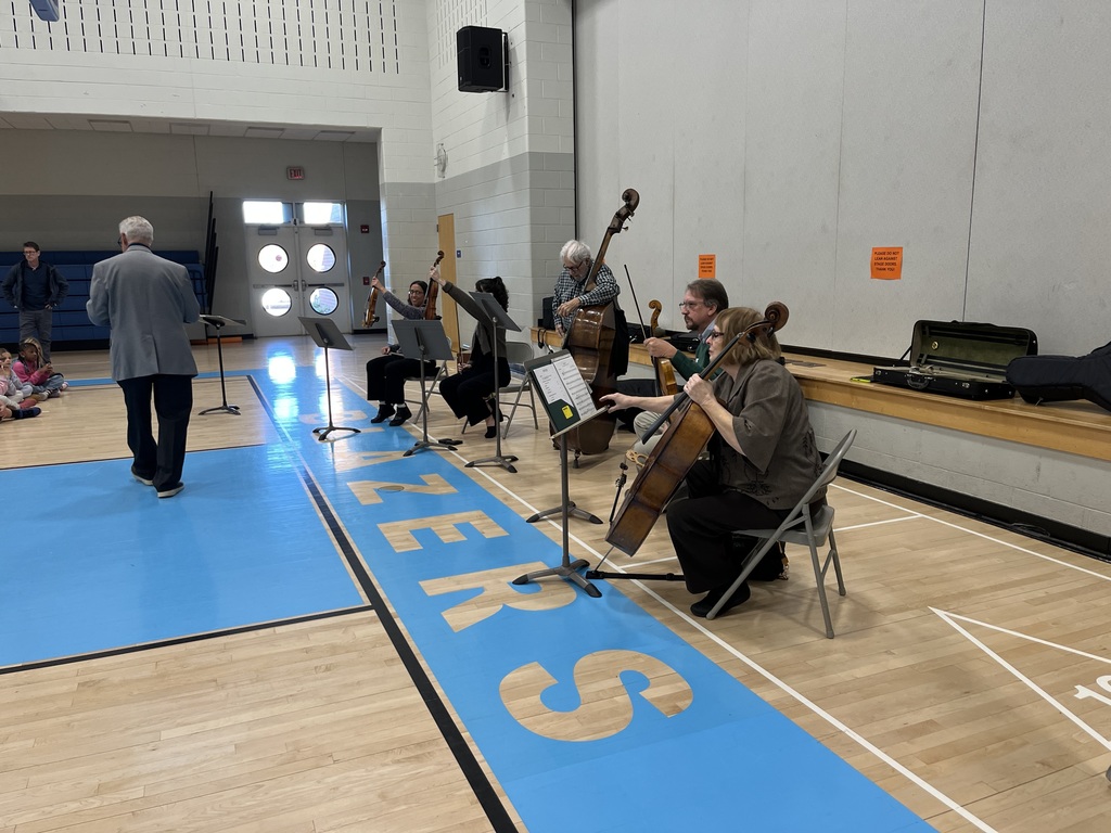 A group of local musicians performs for students in the Intermediate Center gymnasium. The ensemble includes string instruments such as violins, cello, and double bass, seated in front of music stands on the gym floor marked with “BLAZERS.” A presenter stands near the musicians while students sit on the floor watching.