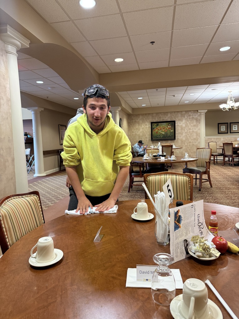 A student in a bright yellow hoodie wipes down a dining table with a cloth. The table is set with cups, utensils, and table number 5. A resident sits in the background, and the dining room features warm lighting and neutral decor.