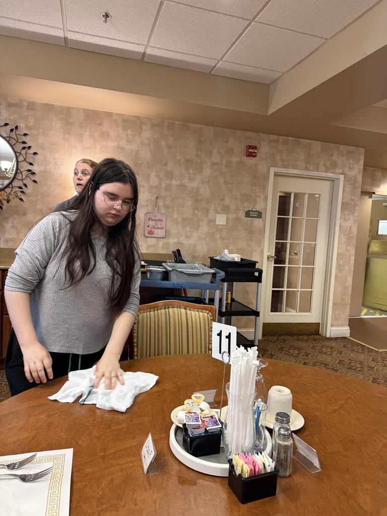 A student wearing glasses and a gray shirt wipes down a dining table with a cloth at a retirement community. Another person is visible in the background near a service cart. Table number 11 and various condiments are on the table.