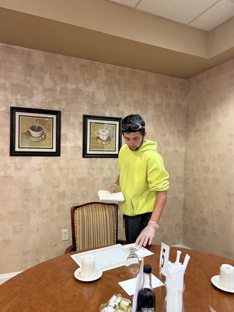 A student wearing a bright yellow hoodie and protective gloves sets a dining table at a retirement community. He is placing a napkin on a placemat while standing beside a wooden table with cups, utensils, and table settings.