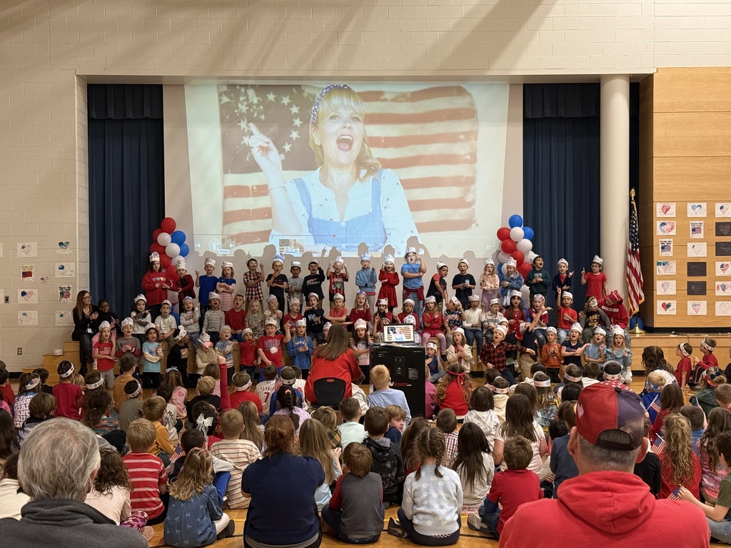 Elementary students wearing patriotic clothing and paper sailor hats perform onstage for a Veterans Day program. A large projected image of a woman singing in front of an American flag appears behind them. Red, white, and blue balloons, student artwork, and seated families fill the gymnasium.