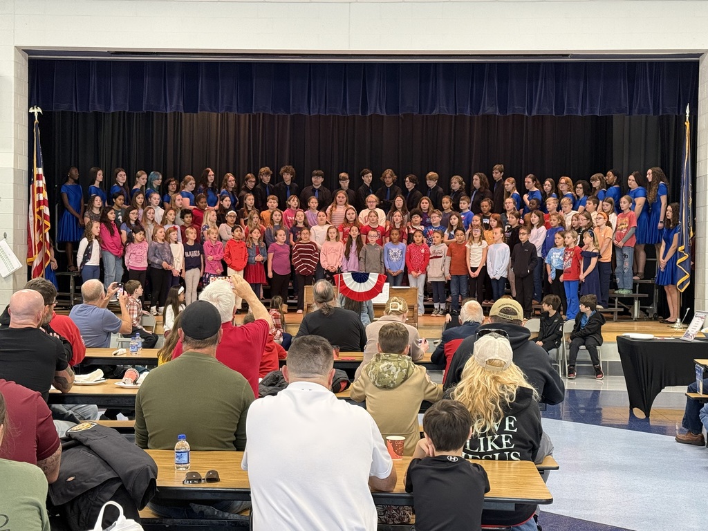 A large group of students from multiple grade levels stand on risers on a stage, dressed in a mix of casual and semi-formal attire, singing for an audience of veterans and families seated at cafeteria-style tables. American flags and patriotic decorations frame the space, creating a Veterans Day celebration atmosphere.