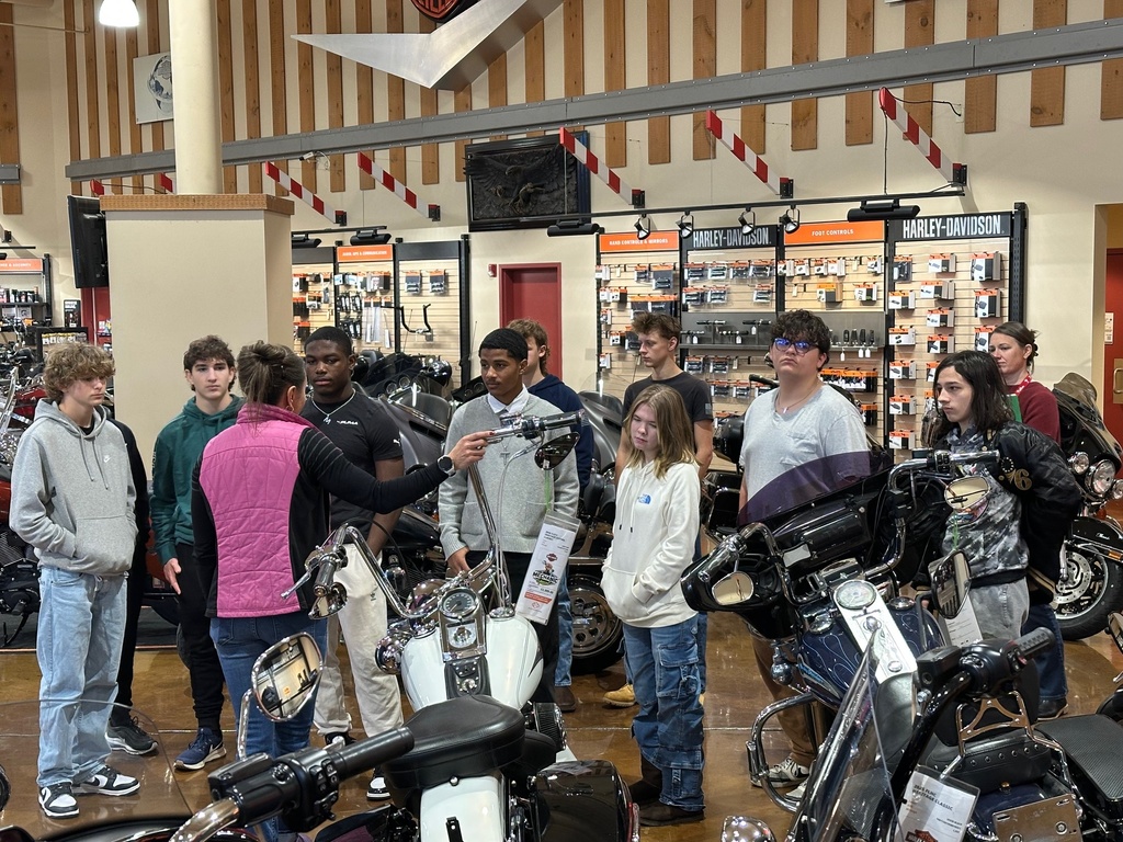 A group of ninth grade students stands around several motorcycles in the main showroom of Classic Harley-Davidson. A staff member gestures toward a bike’s handlebars while explaining features, and students watch attentively among the rows of motorcycles and display racks.