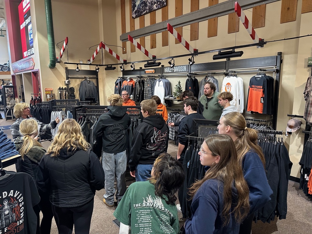 A group of ninth grade students gathers around a clothing display inside the Classic Harley-Davidson apparel section. A staff member speaks to the group while students listen and look at the merchandise.