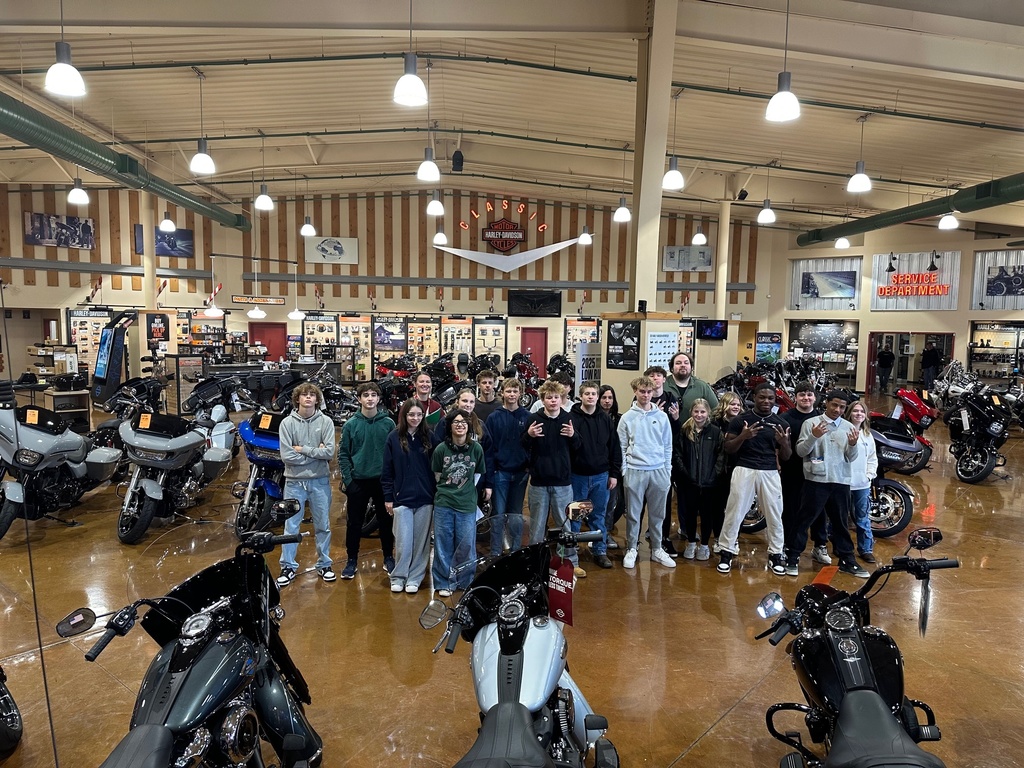 A large group of ninth grade students stands together inside the Classic Harley-Davidson showroom, surrounded by rows of motorcycles. Bright overhead lighting illuminates the space, and the students pose for a group photo among the bikes and showroom displays.