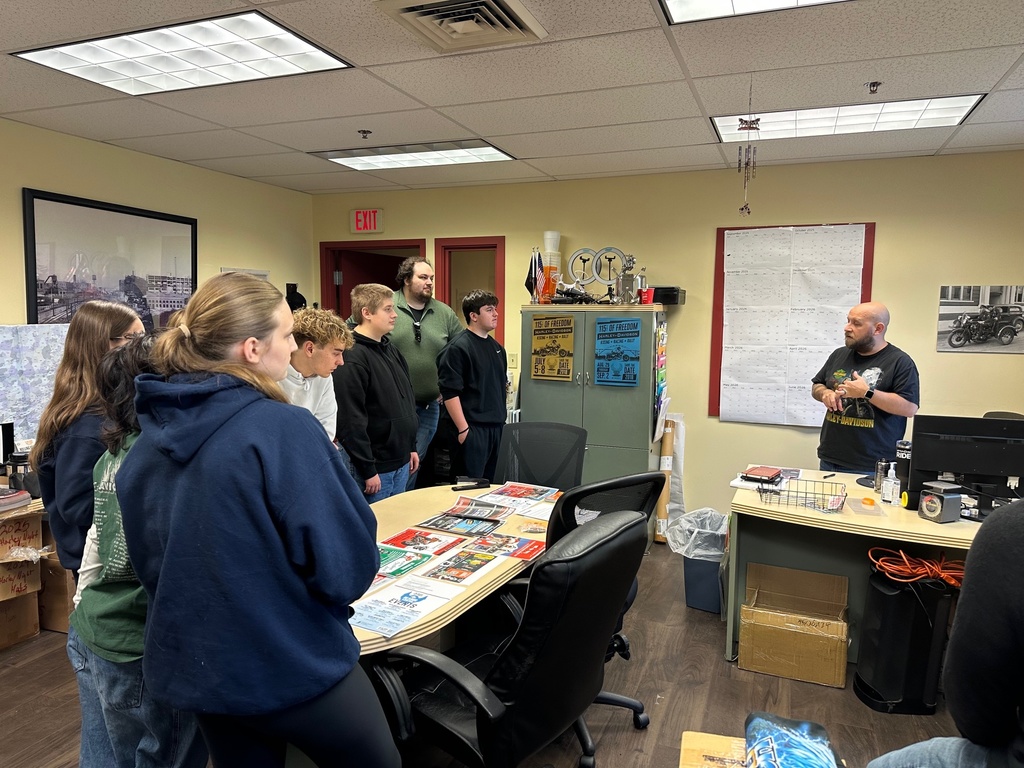 Students stand inside an office at Classic Harley-Davidson, listening to a staff member who is speaking to them. Posters, a wall calendar, and office supplies are visible around the workspace as students observe and ask questions.